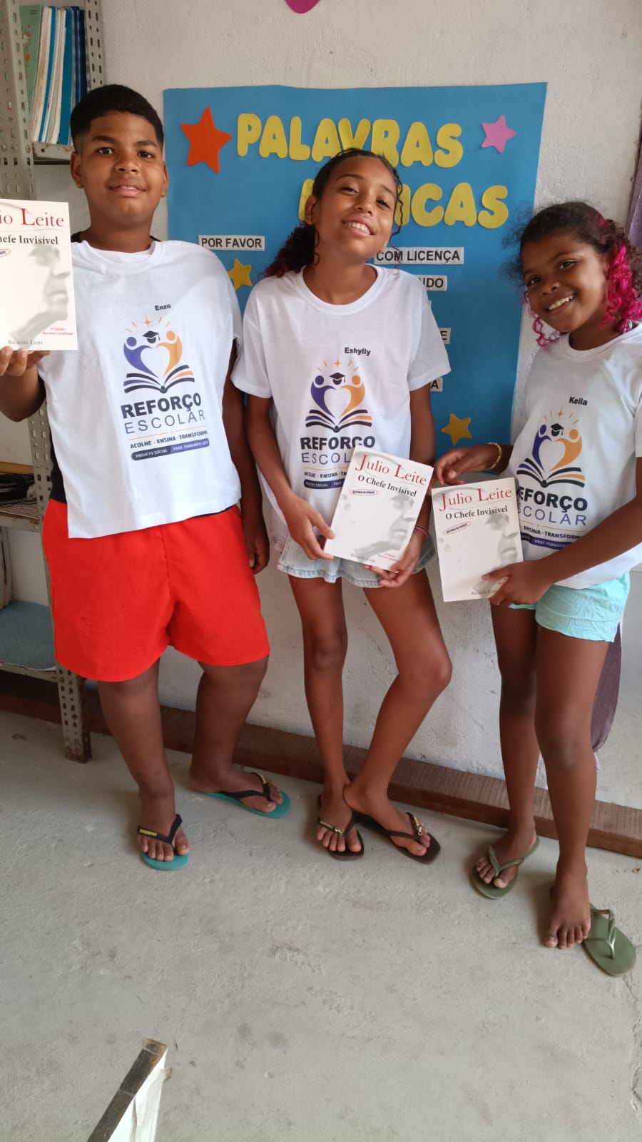 Três alunos do projeto com os livros recebidos, diante do painel das palavras mágicas