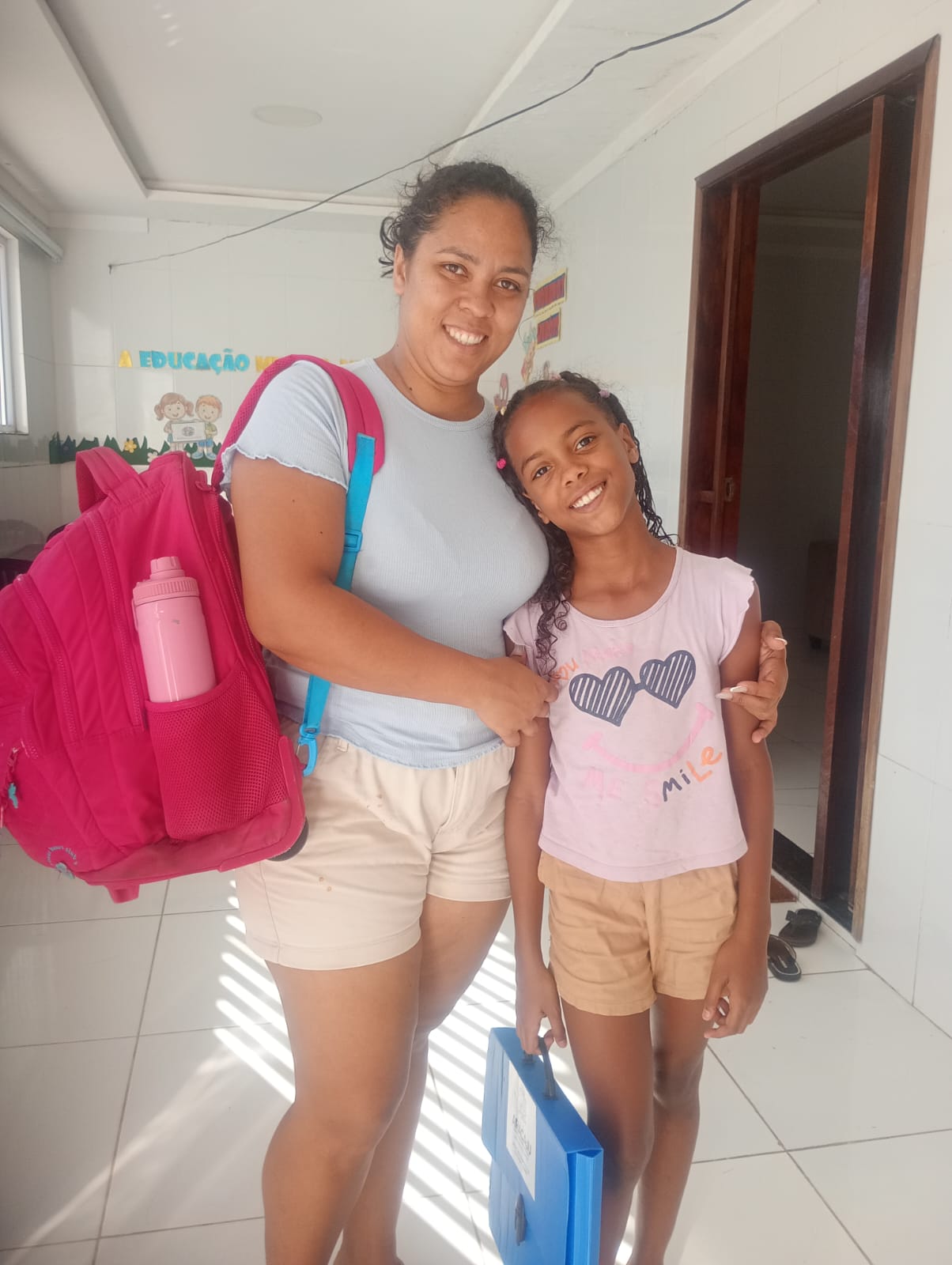 Aluna do projeto ao lado da mãe, com mochila e material escolar